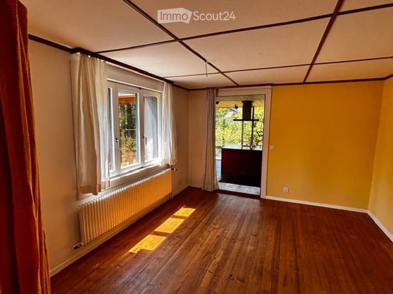 Empty room with wooden flooring, large windows with white curtains, radiator, and a doorway leading to another space.