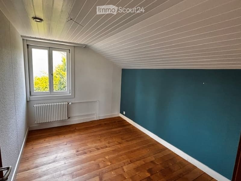 Empty room with wooden flooring, white walls, and a single window with a radiator below it. The ceiling has a slatted design.