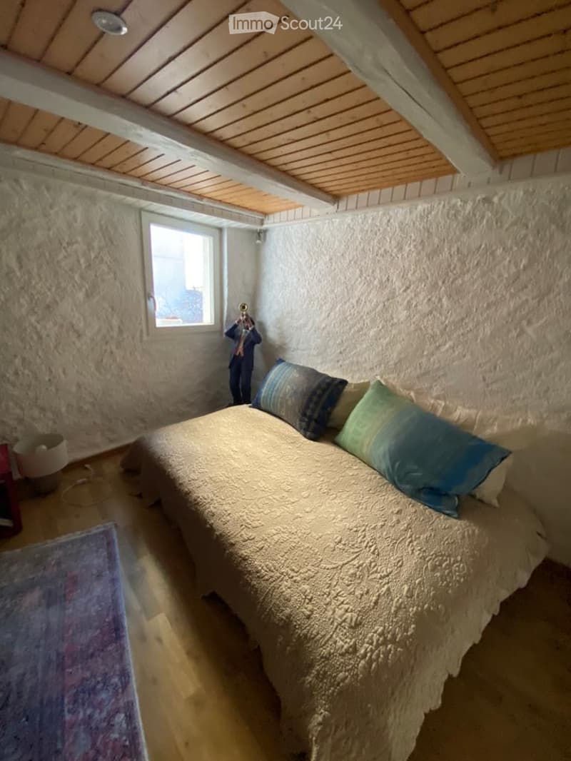 Cozy bedroom with a wooden bed, pillows, a nightstand with a lamp, and a colorful rug on the floor. The walls are textured, and there is a small window letting in natural light.