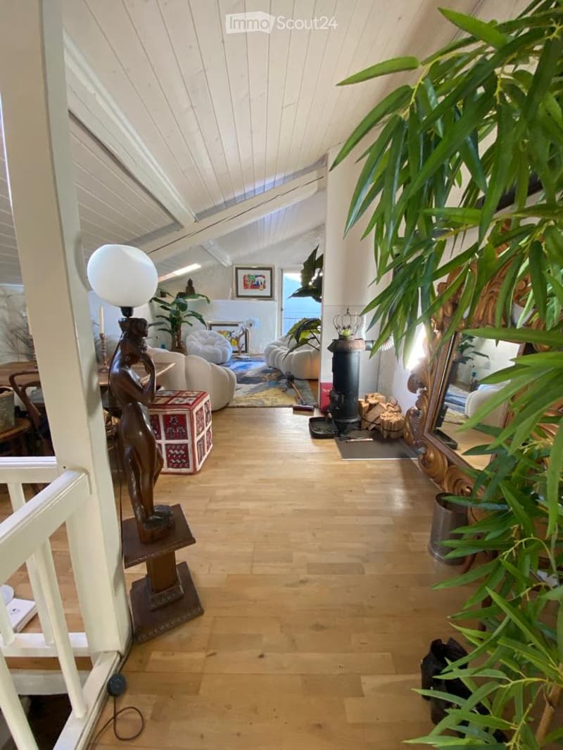 Bright, open living space with wooden floors, white paneled ceiling, fireplace, sofa, chairs, table, lamp, and decorative plants.