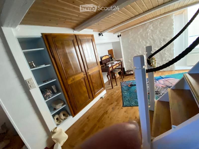 A furnished room with wooden flooring, a large wooden wardrobe, a desk with a chair, a shelf with various items, and a colorful rug.