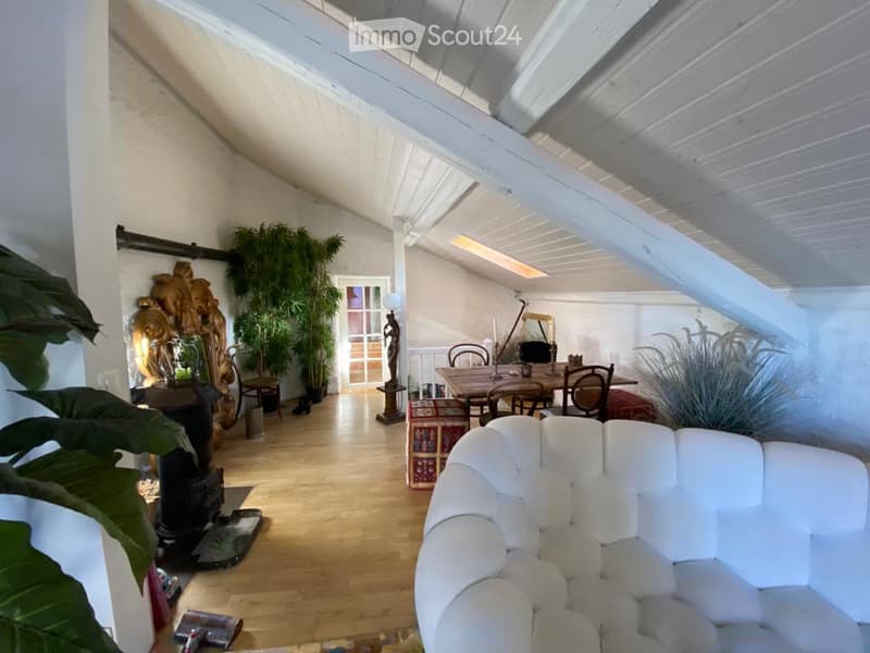 Bright, airy room with wooden flooring, white walls, and exposed wooden beams. Features a white tufted sofa, dining table with chairs, decorative mirror, and several potted plants.