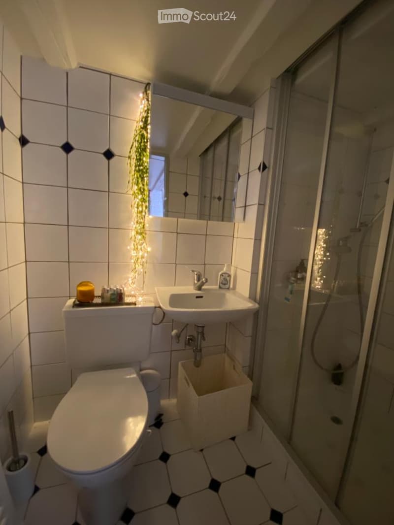 Modern bathroom with white tiles, a toilet, sink, mirror, and a glass-enclosed shower. Features a decorative light string and a small trash bin.