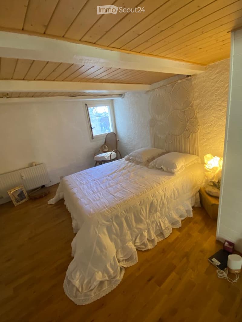 Cozy bedroom with wooden floor, slanted wooden ceiling, white walls, a large bed with white bedding, a nightstand with a lamp, a small chair, and a decorative wall hanging.