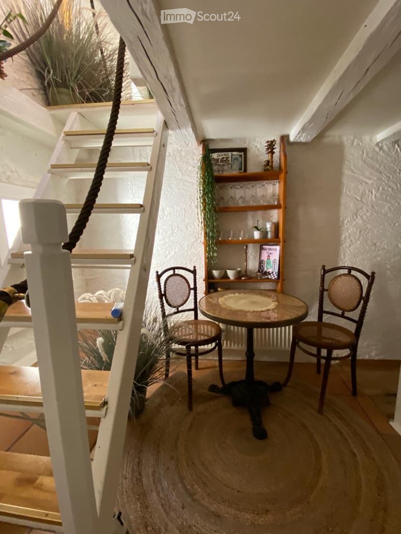 Cozy, well-lit room with a small round table, two chairs, a wooden shelf with decorative items, and a staircase leading upwards.