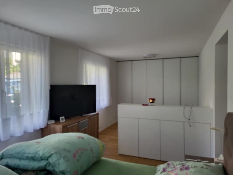 Bright bedroom with a green bed, pillows, a television on a wooden stand, white cabinets, and sheer curtains on the windows.