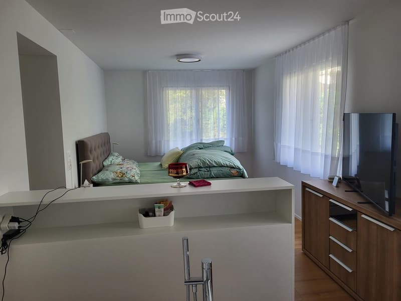 Modern bedroom with a large bed, nightstand, table, chair, and television. The room features white walls, wooden flooring, and large windows with sheer curtains.