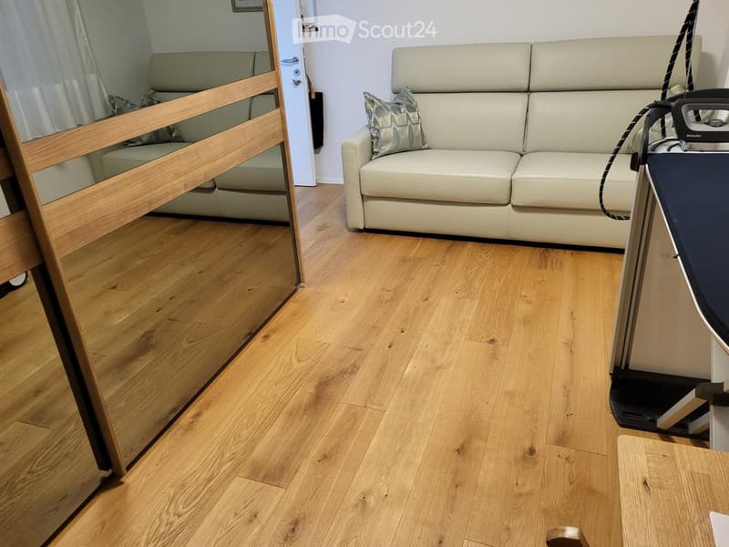 A small, well-lit room with light wooden flooring, featuring a cream-colored sofa with a decorative pillow, an ironing board with an iron, and a small wooden table.