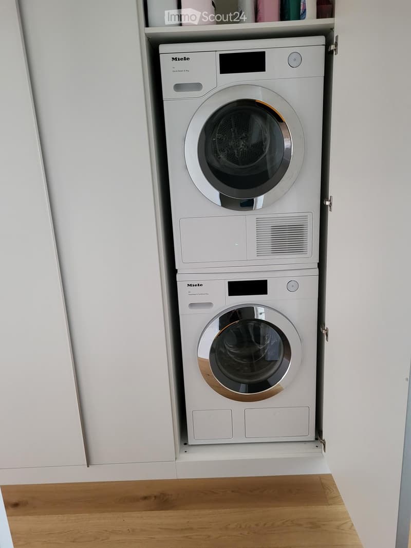 A compact, modern laundry area with a stacked Miele washing machine and dryer unit, white cabinetry, and wooden flooring.