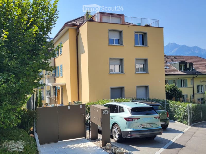 A modern yellow house with multiple windows, a balcony on the top floor, and a gated driveway with a parked car.