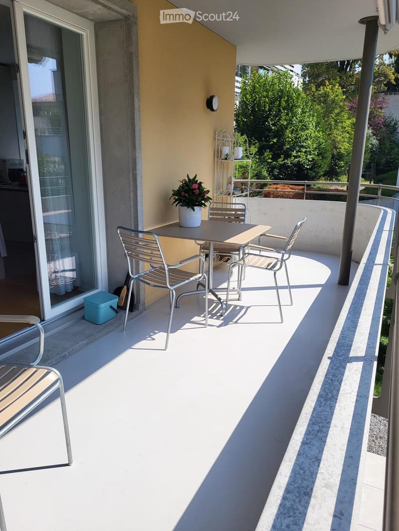 Balcony with a small table, chairs, and a flower pot, overlooking a green area.