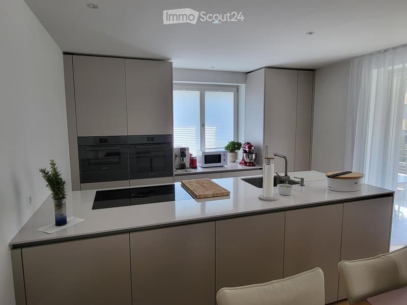 Modern kitchen with grey cabinets, stainless steel appliances, island with sink and stove, and small seating area with chairs.