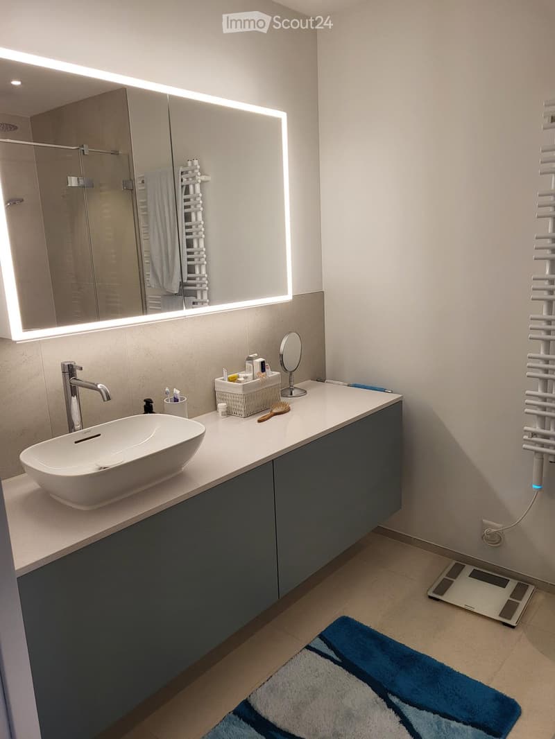 Modern bathroom with a white sink, dark blue cabinetry, large illuminated mirror, towel rack, and a blue and white bath mat on the floor.