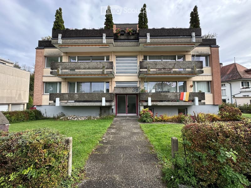 A three-story residential building with balconies, large windows, and a well-maintained garden in front.