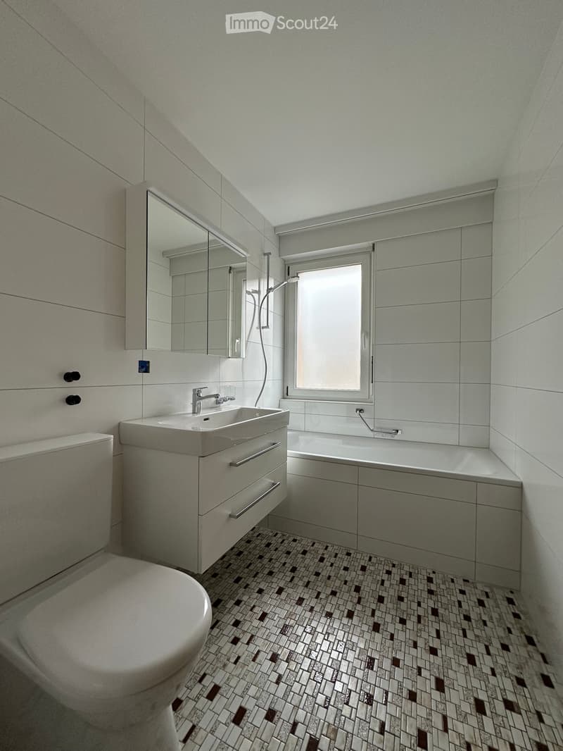 Modern bathroom with a toilet, sink, bathtub, and large mirror. Features a window, sleek cabinetry, and patterned tile flooring.
