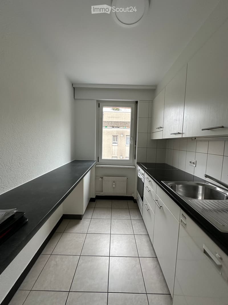 Modern kitchen with white cabinets, black countertops, stainless steel sink, and tiled floor. Features a window with a view of a building outside.