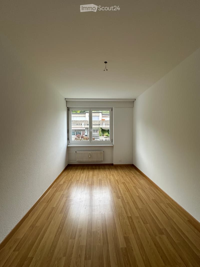 Empty room with wooden flooring, white walls, and a window with blinds. A radiator is visible below the window, and a small light fixture hangs from the ceiling.