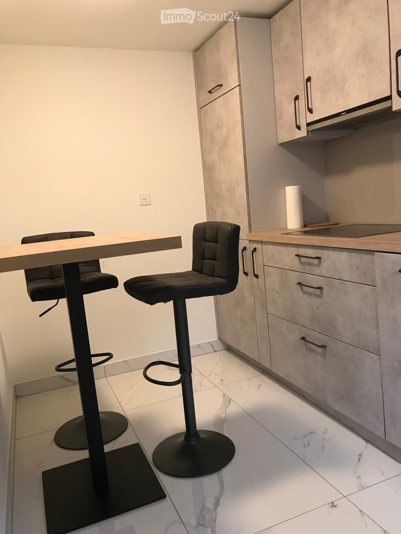 Modern kitchen with light-colored cabinets, a wooden countertop, and two black bar stools near a small table.