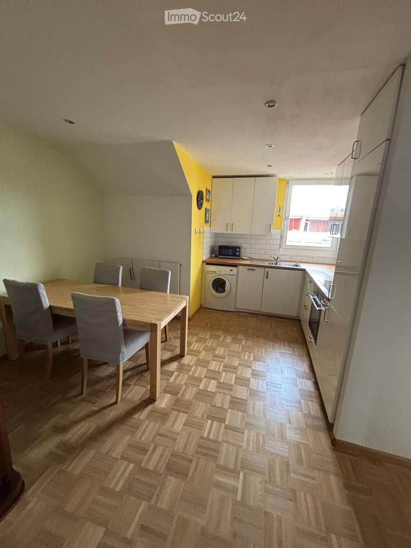 A modern kitchen and dining area with a wooden dining table, four chairs, a microwave, a washing machine, a sink, a faucet, and an oven. The floor is wooden, and the walls are painted in neutral tones with a yellow accent wall.