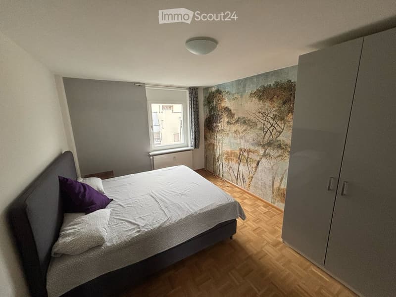 Bedroom with a double bed, pillows, and a large wardrobe. The room features wooden flooring and a scenic mural on one wall.
