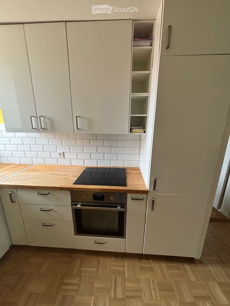 Modern kitchen with light-colored cabinets, wooden countertops, an induction cooktop, and an oven. White tiled backsplash and wooden flooring.