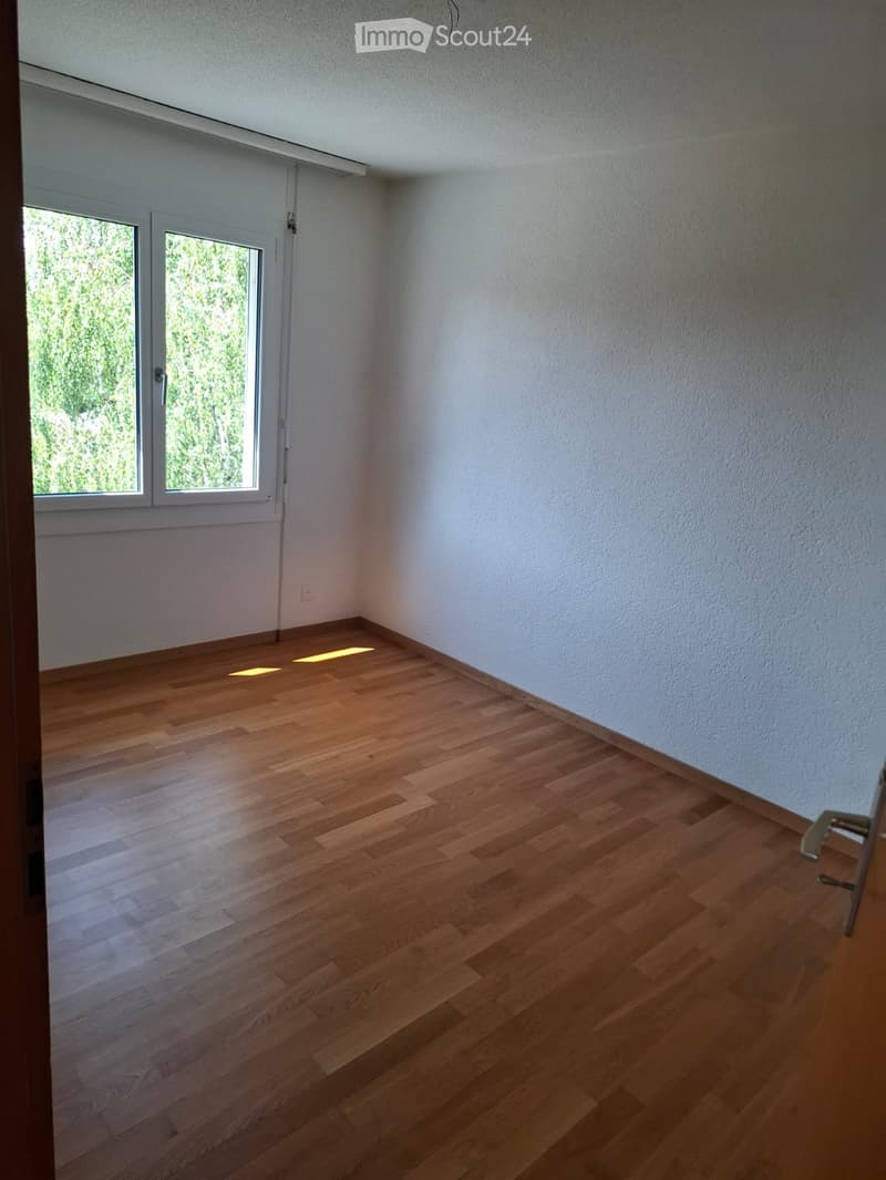 Empty room with wooden flooring and white walls, featuring large windows with a view of greenery outside.