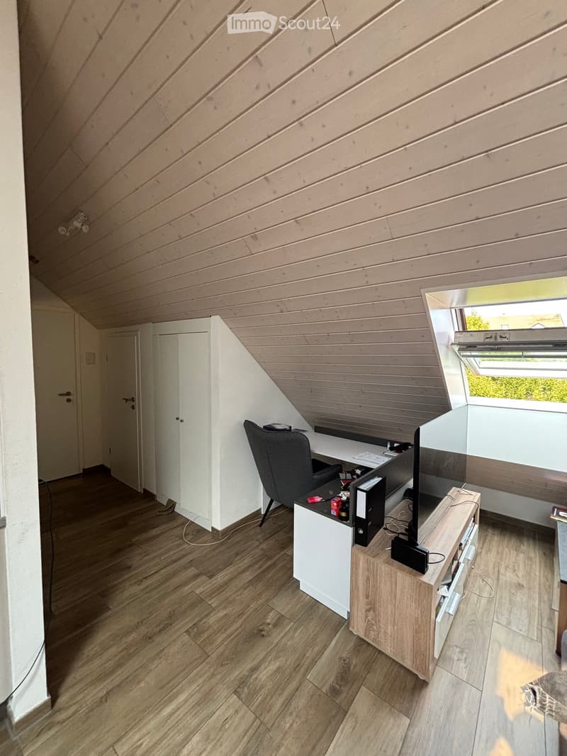 Modern attic room with wooden slatted ceiling, wooden floor, white walls, and built-in desk and cabinet.