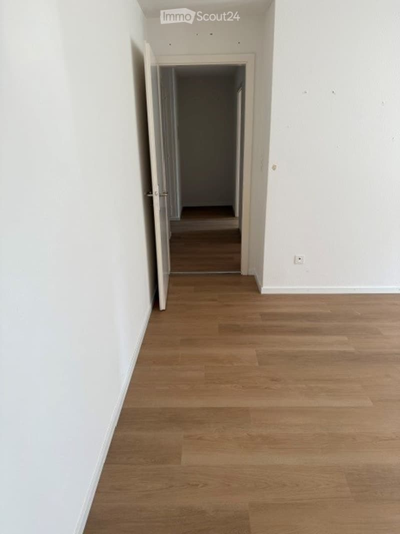 Empty hallway with light wooden flooring and white walls, featuring an open doorway leading to another room.