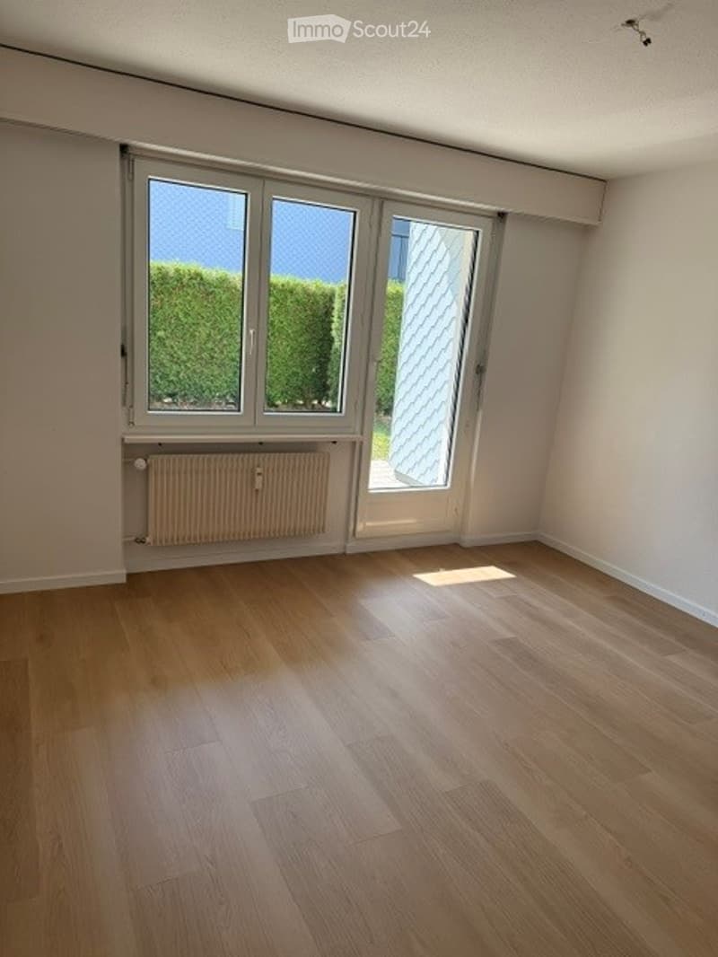 Empty room with wooden flooring, large windows, radiator, and a sliding door leading outside.