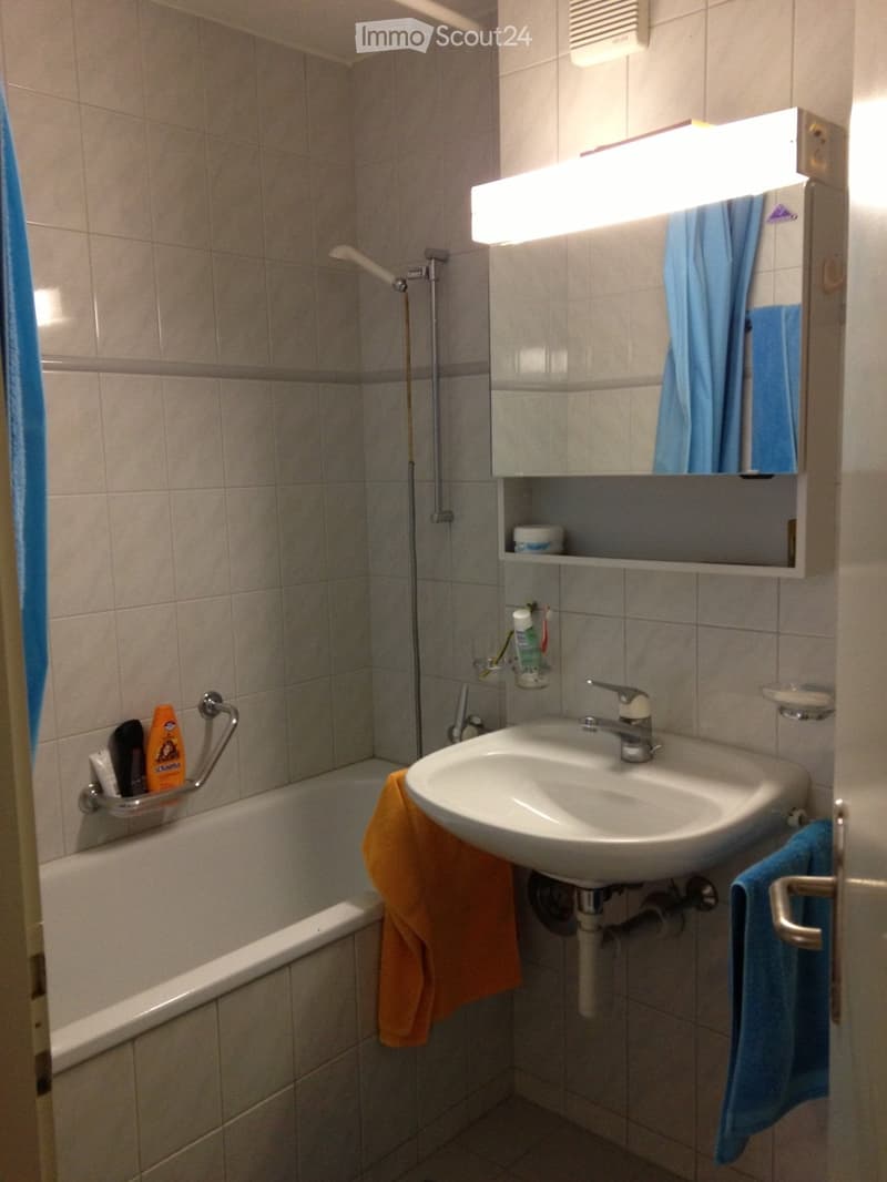 A small bathroom with a bathtub, sink, mirror, and various toiletries. The walls are tiled, and there are towels hanging on the sides.