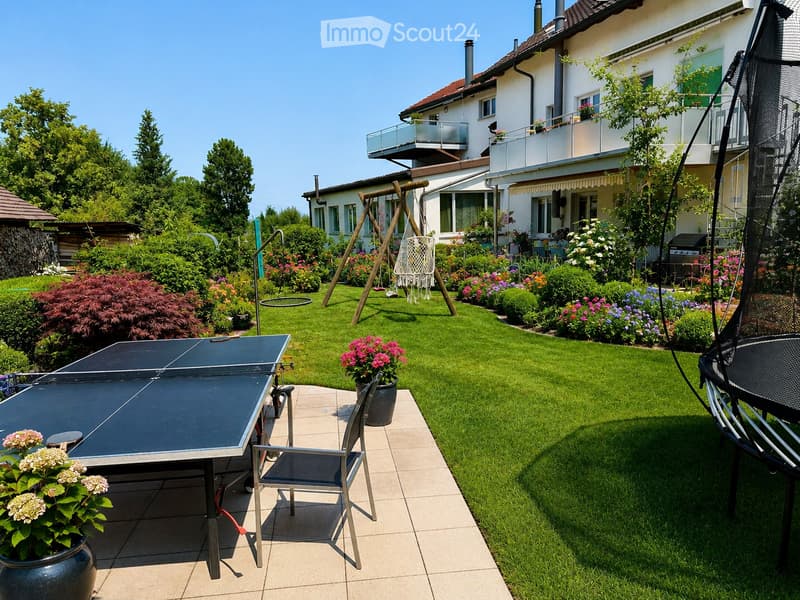 Well-maintained garden with a table tennis table, swing, trampoline, and colorful flower beds surrounding a lawn.