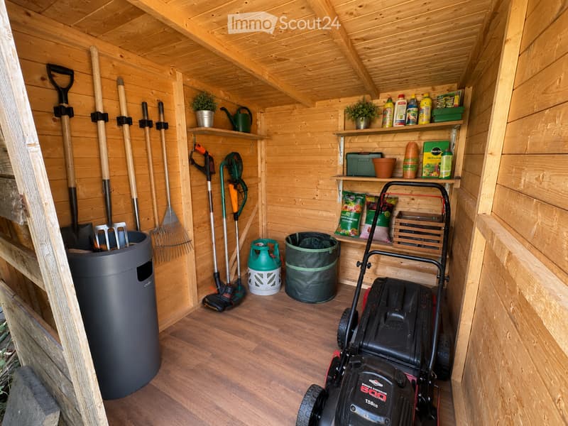 A wooden shed filled with gardening tools, including a lawn mower, shovels, rakes, garden shears, watering cans, potted plants, and storage bins.