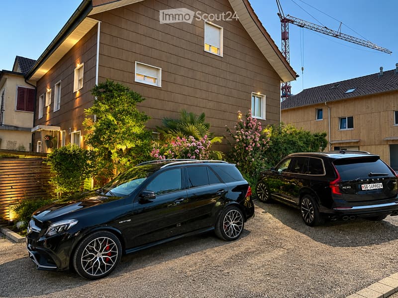 Two-story house with wooden siding, multiple windows, and a well-maintained garden with various plants and flowers. Two black SUVs are parked in front of the house.