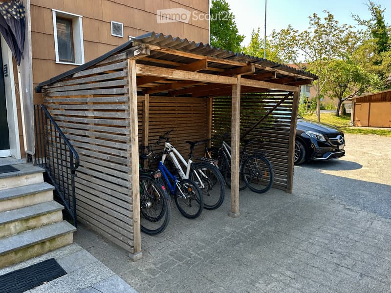 Wooden carport with bicycles parked underneath, adjacent to a house entrance with steps and a railing.