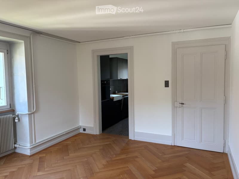 Empty room with wooden herringbone flooring, white walls, and a radiator. A doorway leads to a kitchen area with dark cabinets and a sink.