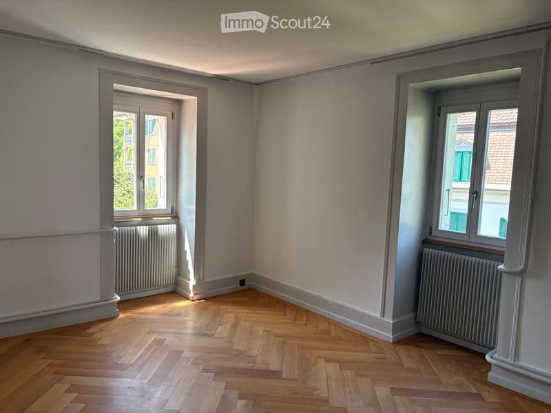 Empty room with wooden parquet flooring, light-colored walls, and two windows with radiators beneath them.