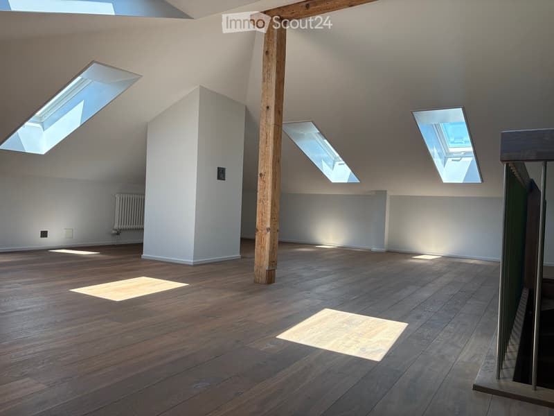 Bright, empty attic space with wooden flooring, white walls, exposed wooden beam, and multiple skylights allowing natural light to flood the room.