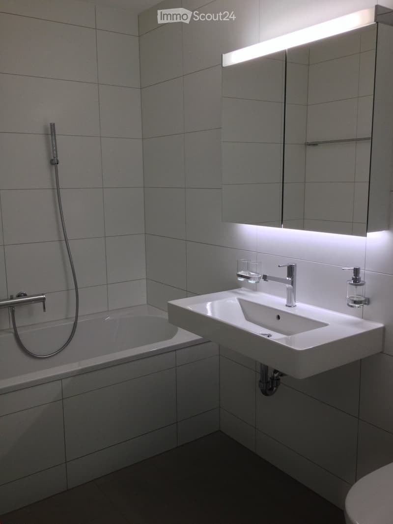 Modern bathroom with a white tiled wall, a bathtub, a sink with a modern faucet, a soap dispenser, and a large mirror with integrated lighting.
