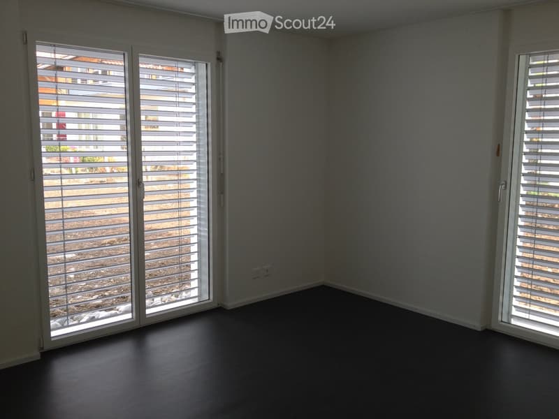 Empty room with dark flooring, white walls, and large windows with blinds overlooking an outdoor area.