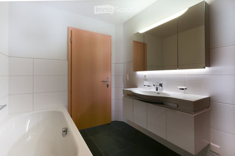 Modern bathroom with a white bathtub, a sink with a modern faucet, a large mirror with built-in lighting, and a wooden door.