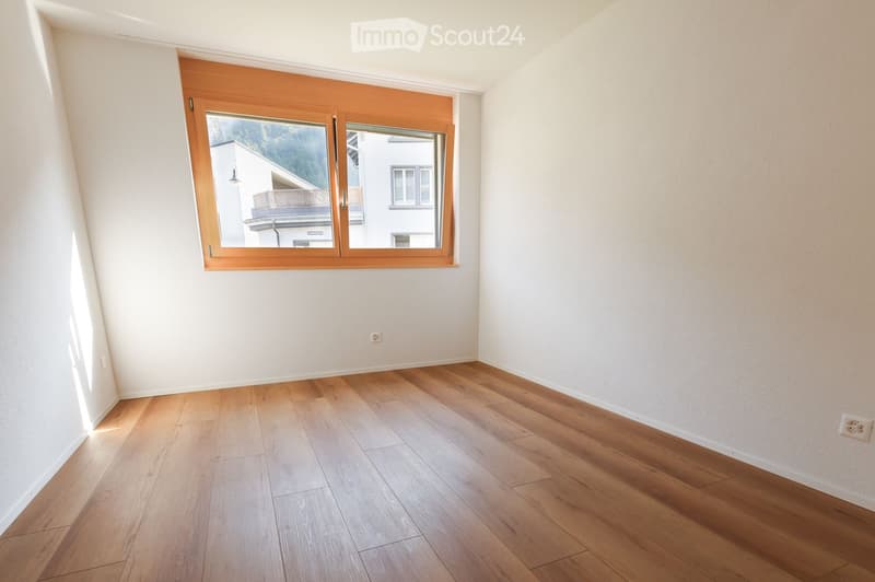 Empty room with light wooden flooring and white walls, featuring a large window with wooden frame and blinds.