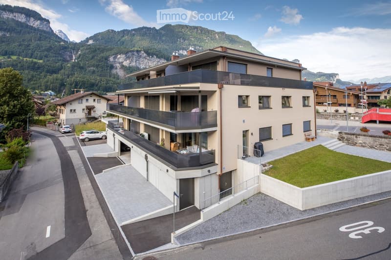 Modern multi-story building with balconies, surrounded by a parking area and scenic mountain backdrop.