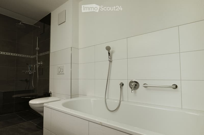 Modern bathroom with a white bathtub, toilet, and a glass-enclosed shower area with a handheld showerhead. The walls are tiled in white and black, and there is a handrail next to the bathtub.