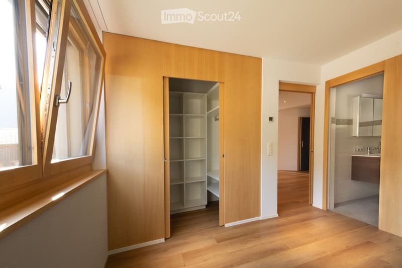 Empty room with wooden flooring, large window with wooden frame, built-in closet with empty shelves, and a view of a bathroom area with sink and cabinets.