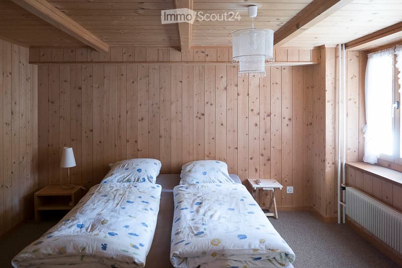 Cozy wooden bedroom with two single beds, matching bedding, nightstands, lamps, and a small table with a remote control. Wooden walls and ceiling beams, large window with white curtains.