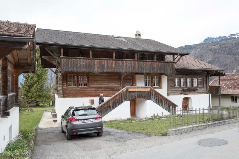Traditional wooden house with a balcony, multiple windows, and a staircase leading to the entrance. A car is parked in front, and the house is surrounded by greenery and a mountainous backdrop.