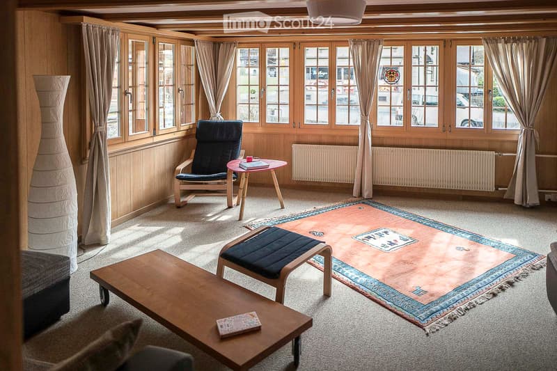 Cozy living room with wooden walls, large windows, a sofa, coffee table, chairs, a small table, and a decorative rug.