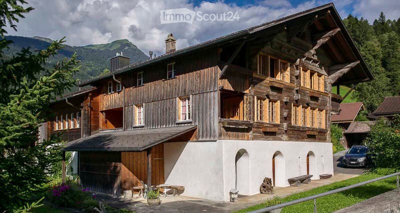Traditional wooden chalet with multiple stories, balconies, and a garage, set against a mountain backdrop.