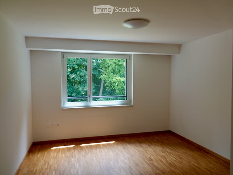 Empty room with wooden flooring, white walls, and a large window showing greenery outside. No furniture or equipment present.