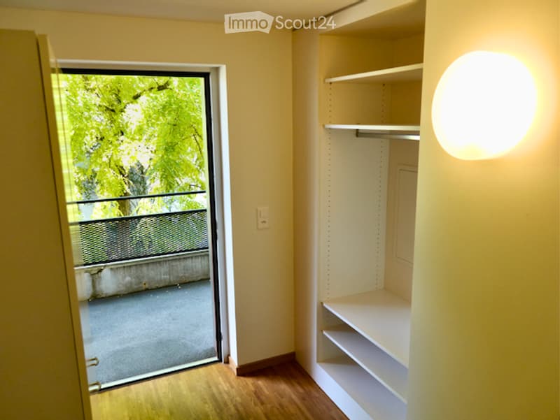 Empty room with wooden flooring, built-in shelving unit, and a large window with a view of greenery outside.
